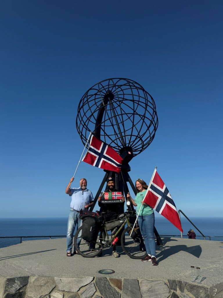 Tre mennesker med norske flagg og en sykkel foran et monument.