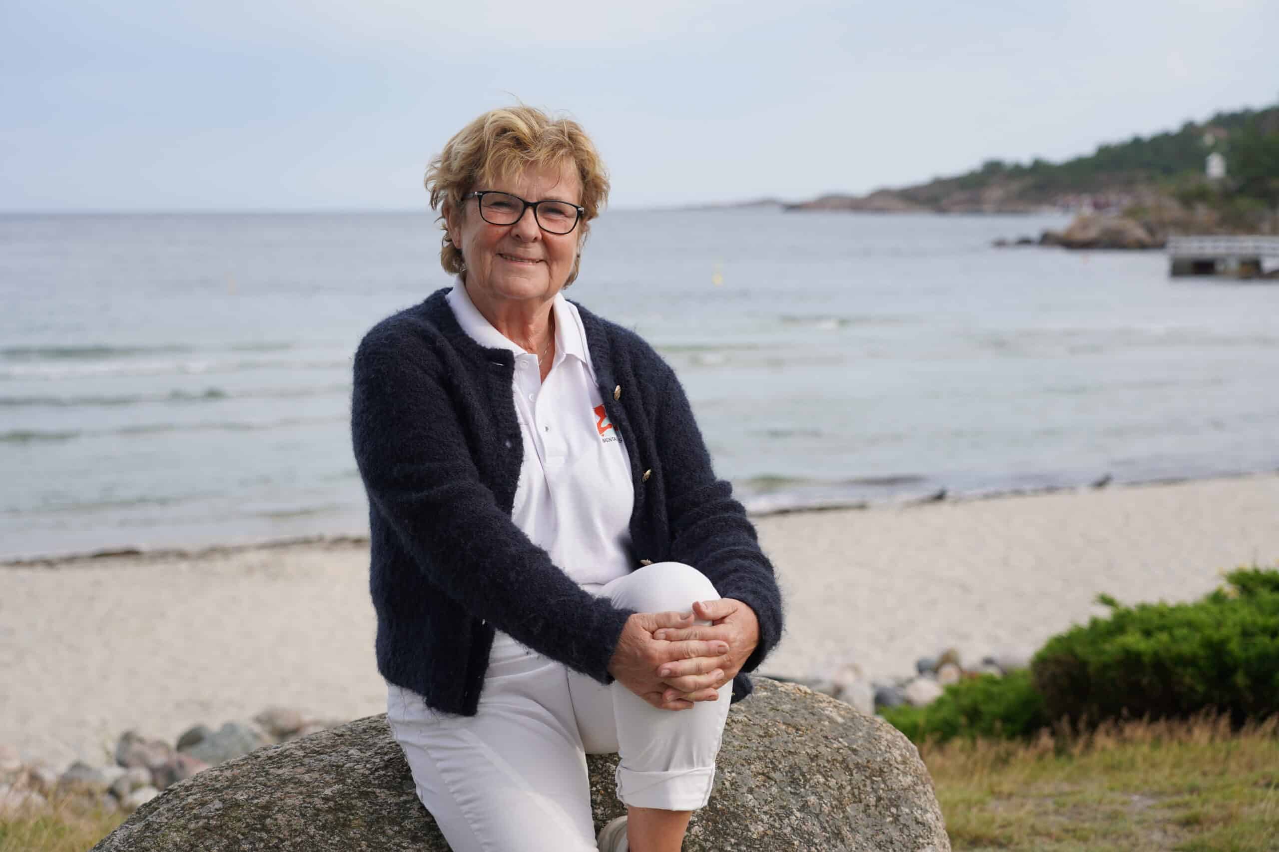 Aslaug Timland Dale daglig leder på Hjelpetelefonen smiler sitter på en stein med utsikt over sjøen bak seg.