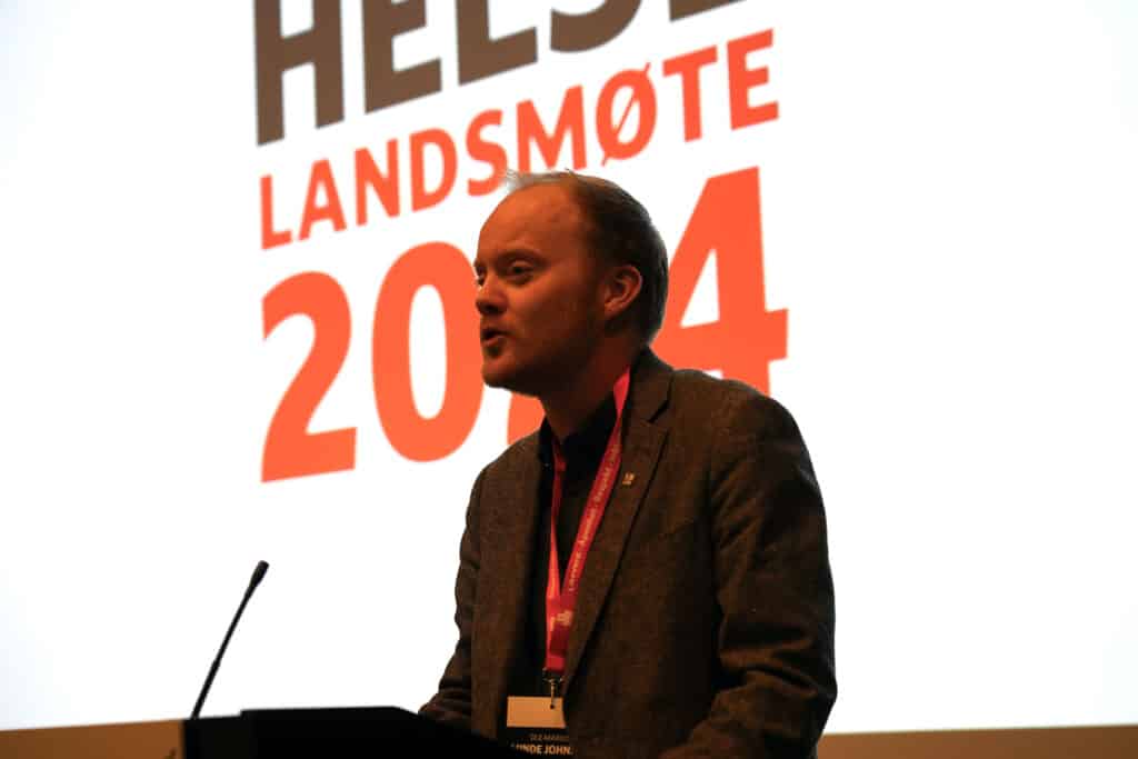 Mann snakker i mikrofon foran stor skjerm.