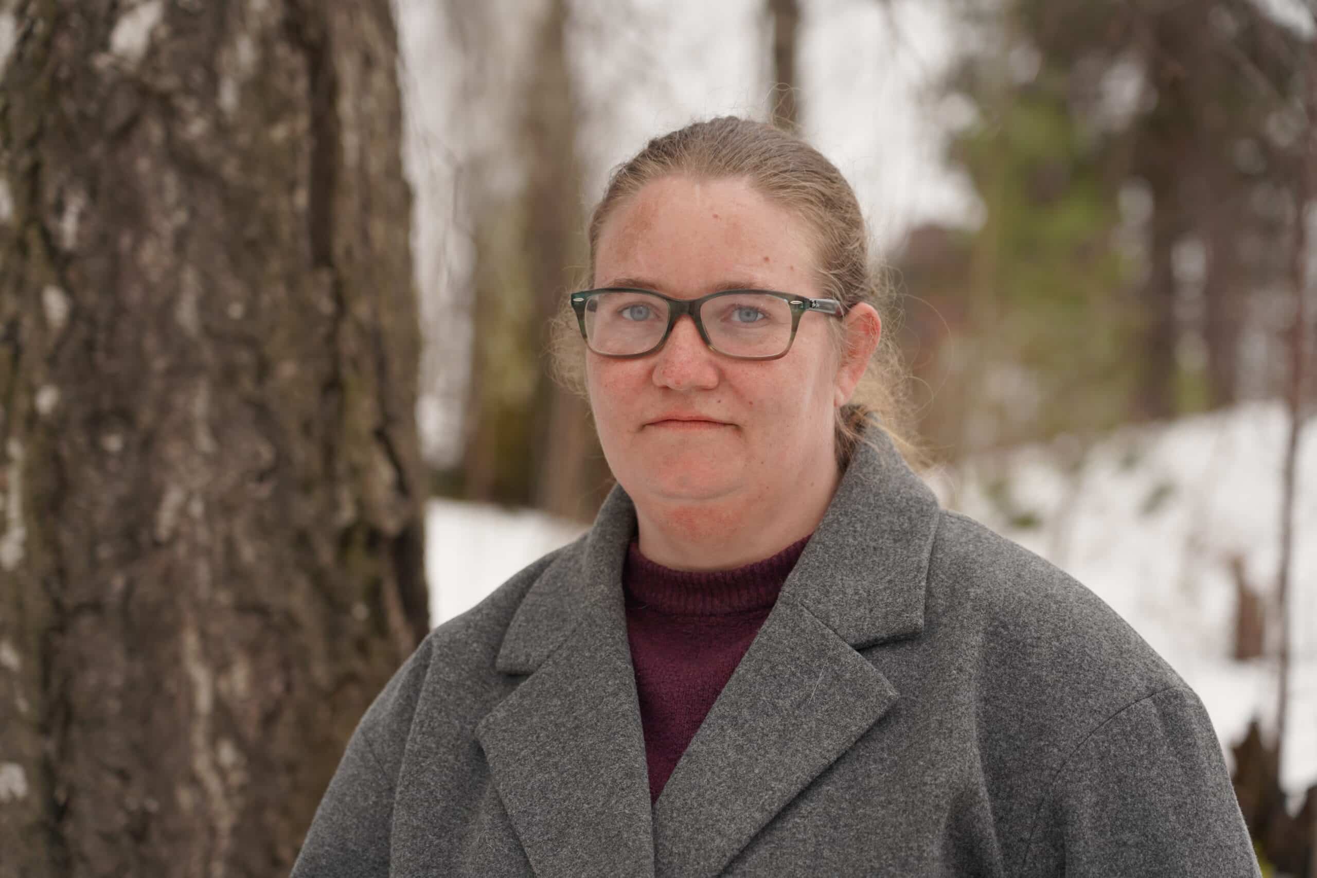 kvinne i grå jakke står ute i skogen og ser alvorlig inn i kameraet.